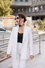 Fototapeta premium Stylish woman in a striped suit confidently walking outdoors with sunglasses and chic hairstyle