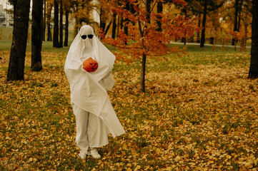 Woman in white sheet holding jack o'lantern against autumn forest background. Halloween costume. 
