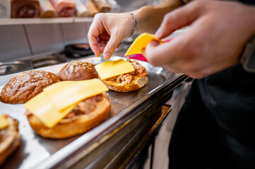 A close-up of hands placing cheese slices on burger buns with cooked patties, suggesting food preparation in a kitchen setting. Ideal for culinary, cooking, and food-related themes.