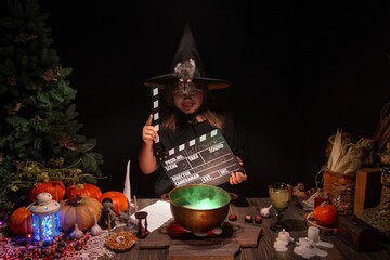 Halloween, smiling little cute girl with Film Clapperboard, dressed as witch in witch's hat brews a glowing magic potion. Magic wand, сandles, pumpkins, fog smoke, magic, mystical. Witch and rat pet.