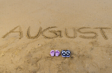 Hand written august month baby announcement text is on the beach sand. Clean water and small waves are in background