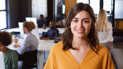 Portrait Of Smiling Young Businesswoman Working In Busy Modern Office With Colleagues In Background