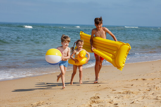 Children run along beach, carrying inflatables and a beachball - Powered by Adobe