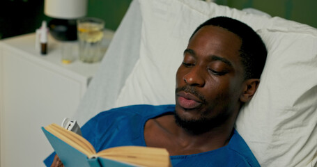 In the hospital room, the guy is lying on a bed, finding solace in reading during his treatment.