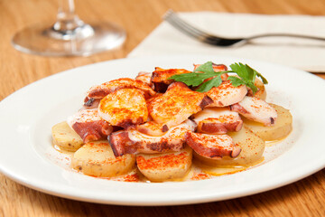 Octopus dish with sliced ​​potatoes with paprika on a white plate with fork, napkin and glass of wine on a wooden background