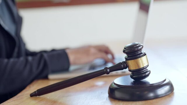 A person is using a laptop computer while a gavel sits on a table in front of them. The person appears to be working on a legal document or case, as the gavel is a symbol of authority and justice