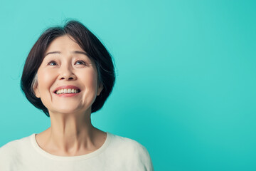 Smiling elderly Asian woman looking upwards against a bright turquoise background.