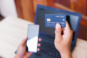 A person holding a credit card in one hand and a smartphone in the other while near a laptop.