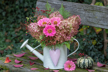 Blumenstrauß mit pink Zinnien, Rispenhortensien-Blüten und Hagebutten in dekorativer Gießkanne auf Gartenbank