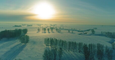 Drone aerial view of cold winter landscape arctic field, trees covered with frost snow, ice river and sun rays over horizon. Extreme low temperature weather.