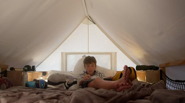 Caucasian boy sitting in a tent, focused on reading and writing in a notebook. Various objects like a ball and a virtual reality headset are scattered around him inside the tent