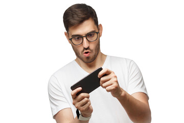 Young man in white t-shirt holding phone with both hands, moving it while playing video game in app, making funny face