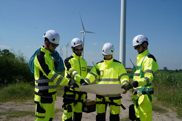 Fototapeta premium A group of engineers wearing reflective forms hold blueprints to inspect the installation of wind turbines to generate electricity in a windmill field.... 