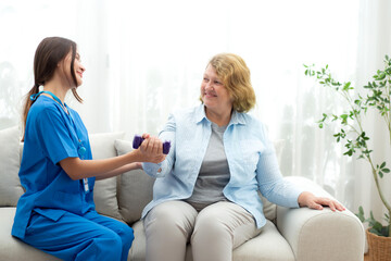 Fototapeta premium Asian caregiver woman or nurse training senior woman lifting dumbbell for arm exercise while therapy and rehabilitation in living room at home, caretaker or physiotherapist helping elderly workout.