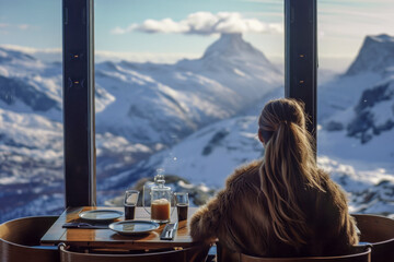 A woman sits at a table in a restaurant with a view of mountains. She is wearing a fur coat and has her hair in a ponytail