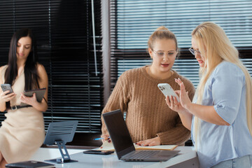 Two women stand at the table and discuss something while looking at the phone, the third woman...
