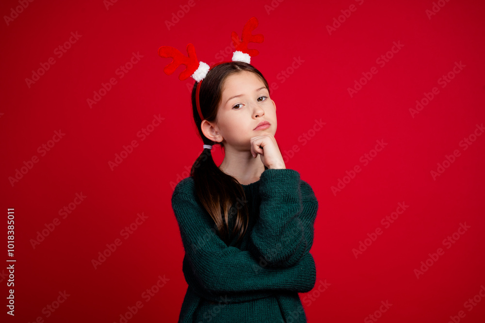 Wall mural Photo portrait of cute little girl thoughtful touch chin dressed green christmas clothes theme party on red color background