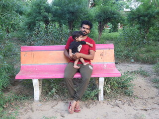 A handsome father sits with his 1-year-old baby girl on a cement seat amidst greenery in Govardhan...
