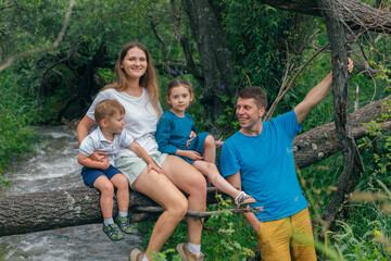 Fototapeta premium A joyful family is bonding together on a fallen tree by the flowing river in a beautifully lush green environment