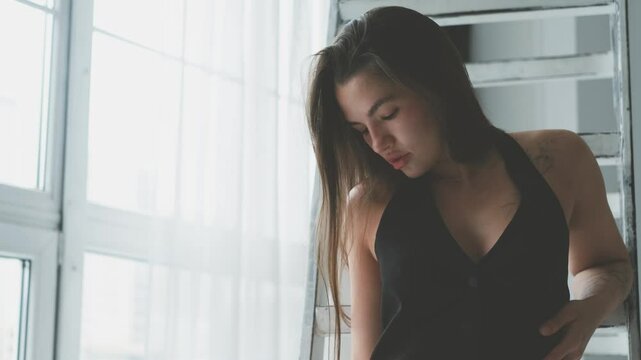 Portrait of young beautiful model posing in black lingerie and stockings near a large window. Calm cute brunette in fashionable lingerie.