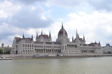 Obraz premium A daytime view of the Hungarian Parliament Building from a boat on the Danube River. The stunning Gothic Revival structure rises majestically, its spires and domes reflecting in the water,