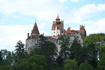 Fototapeta premium The photo captures Bran Castle, perched dramatically atop a rocky hill. The medieval fortress, with its red-tiled roofs and stone walls, stands against a backdrop of lush green trees.