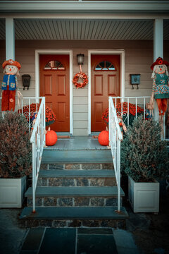 Two bright orange doors are surrounded by autumn decorations, including scarecrows, pumpkins, and red flowers. A set of stairs leads up to the porch, which is decorated for the fall season.