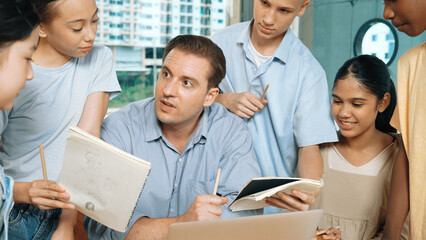 Obraz premium Caucasian teacher giving comment or advise student while smart bot pointing at laptop screen at classroom. Diverse student taking a note while teacher teach lesson while using computer. Edification.