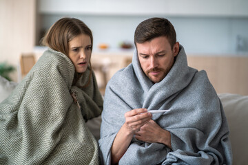 Weak shivering family wrapped in blanket sitting on sofa, man measuring temperature with...