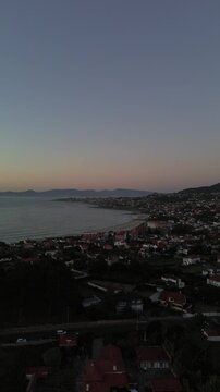 Aerial view of Patos and Prado beaches with houses above, after sunset. Toralla island and Vigo estuary are visible in the distance. Monteferro obscures the view as camera descends to Area Fofa beach.