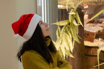 a woman with dark hair and a Santa Claus hat looks dreamily at a store window