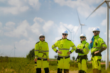 A team of engineers in high-visibility safety gear stand together at a wind farm, conducting inspections and discussing renewable energy projects. Wind turbines surround them under a clear sky.