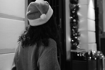 a woman in a santa claus hat walks forward past a house with garlands