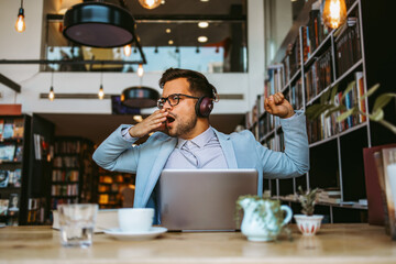 Middle age handsome man sitting in modern bookstore, reading and using his laptop or notebook computer while listening to music with headphones.