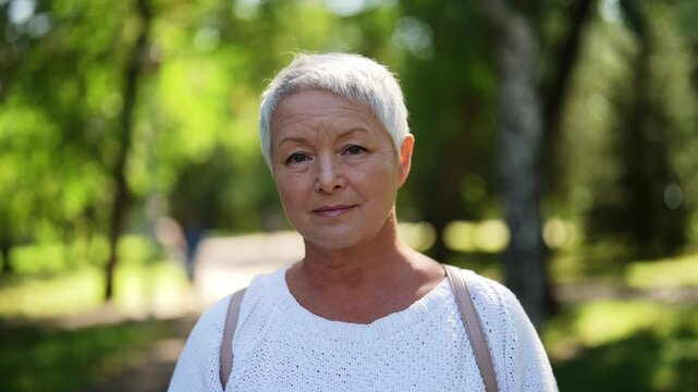 Portrait of happy gray-haired senior woman smiling looking at camera standing posing in green summer park on sunny day, exuding confidence and contentment in stylish casual outfit, enjoying retirement