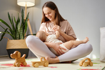 Brown-haired woman sitting on floor in living room lovingly breastfeeding her infant son embracing...