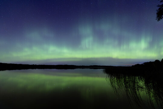Northern Lights erupt over a Minnesota lake in the dark sky overhead shining a bright rainbow of Aurora light and colors over the forests and clean water during strong solar storm