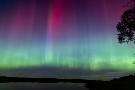 Northern Lights erupt over a Minnesota lake in the dark sky overhead shining a bright rainbow of Aurora light and colors over the forests and clean water during strong solar storm