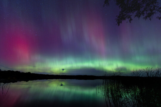 Northern Lights erupt over a Minnesota lake in the dark sky overhead shining a bright rainbow of Aurora light and colors over the forests and clean water during strong solar storm