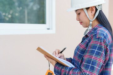 Asian female construction worker inspects new house windows, taking notes on clipboard, focusing on...