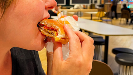 A Caucasian woman enjoys a juicy burger in a vibrant food court, illustrating fast food culture and...