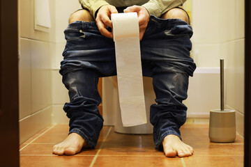 Man Sitting on Toilet Holding Toilet Paper in Bathroom with Pants Down