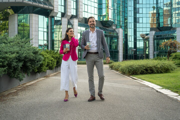 Fototapeta premium Business people walking with coffee in front of glass building. Happy excited office colleagues in stylish outfits talking about work when walking over street in summer. 
