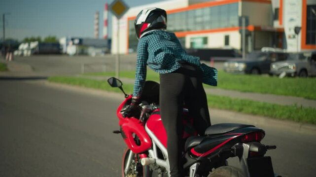 A female motorcyclist, wearing a checkered shirt and canvas shoes, rides a power bike standing, a blue truck is visible ahead of her with parked cars in the background on an open road