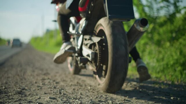 Close-up of a biker kickstarting their motorcycle on the roadside, with a car in the distance approaching in the opposite direction