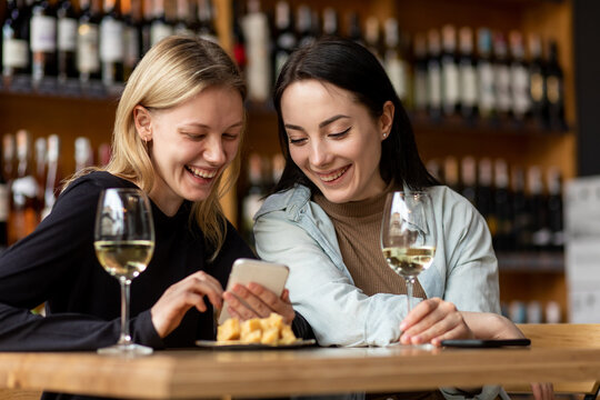 Smiling women looking photos from phone with glasses of wine