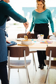 Host Serving Food At Dining Table With Woman In Teal Sweater Nearby