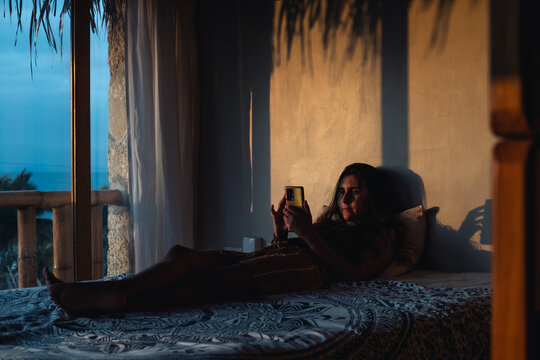 young woman in bed with phone in dark room at dawn