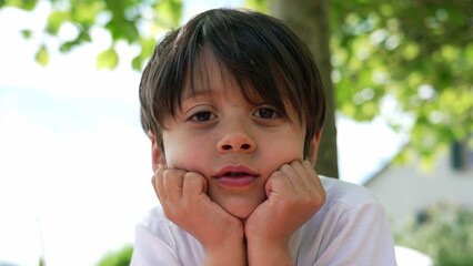 Young boy resting his chin in his hands, looking directly at the camera with a thoughtful expression, seated at an outdoor cafe on a sunny day © Marco