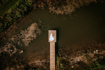 A white wedding dress and shoes are placed on a wooden dock over a murky pond surrounded by greenery, captured from above with ample copy space..